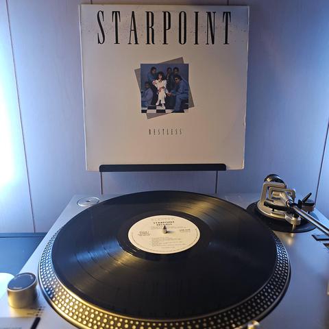 A vinyl record sits on a turntable. Behind the turntable, a vinyl album outer sleeve is displayed. The front cover shows  mostly blank space with a small square image of the members of Starpoint grouped together.
