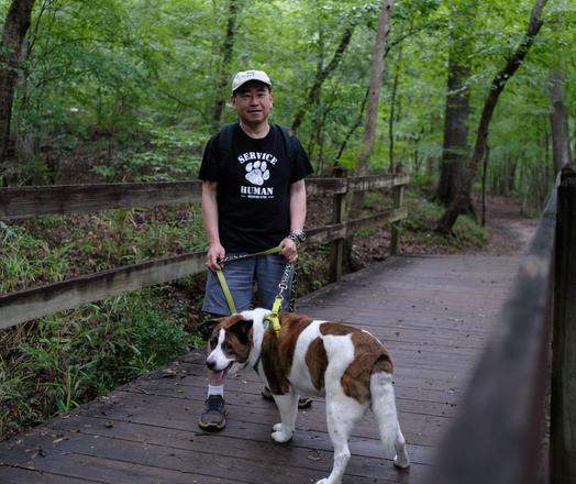 A man with a black T-shirt saying "Service Human" standing on a wooden bridge in a park, holding his white and brown dog