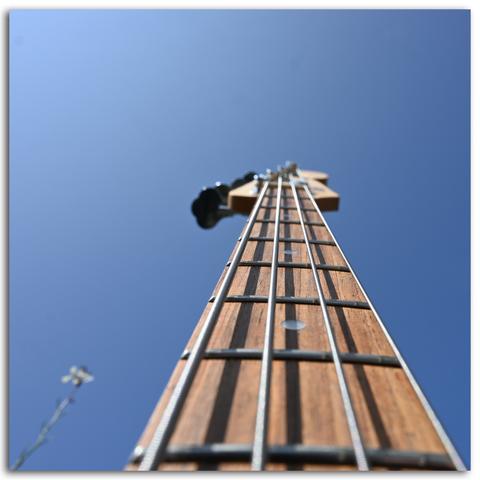Der Hals eines E-Bass reicht in den wolkenlos blauen Sommerhimmel, links daneben ragt im Bokeh ein dünner Stengel einer Balkonpflanze nach oben.