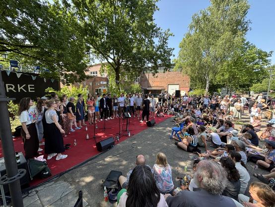 Links der Chor auf einem roten Teppich unter Bäumen rechts das Publikum teilweise auf dem Boden sitzend. Strahlender Sonnenschein.