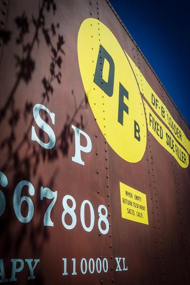 The image shows a close-up view of a large, rust-colored train car. The car's surface is marked with various texts and symbols. Prominently, a large yellow circle with the letters "DFB" in black is visible. Below this, the text "SP" is written in large white letters, followed by the number "67808" in white. A smaller yellow rectangle contains the text "DF-B LOADER FIXED SIDE FILLER" in black. Another yellow rectangle at the bottom right corner reads "WHEN EMPTY RETURN TO SP AGENT SACATO. CALIF." in black. The bottom of the car features the text "CAPACITY 110000 XL" in white. The background is a clear blue sky, and shadows of tree branches are cast on the car's surface, adding depth to the image.

Provided by @altbot, generated privately and locally using Ovis2-8B