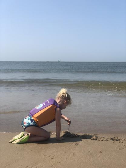 A portrait oriented photo of a blonde 5 year old sitting on her knees in the sand next to the sea, wearing a blue bathing suit, purple and orange swimming vest and neon yellow water shoes. She’s dripping mud through her fingers onto the beach. Her face is not visible, as we find our child’s privacy very important. The sky is blue without clouds, the sea is quite calm with just the start of a small wave starting to grow a few meters from the beach. It was a hot day, luckily there was a nice breeze on the beach. Our kid rescued a lot of jellyfish. She calls herself a nature witch.