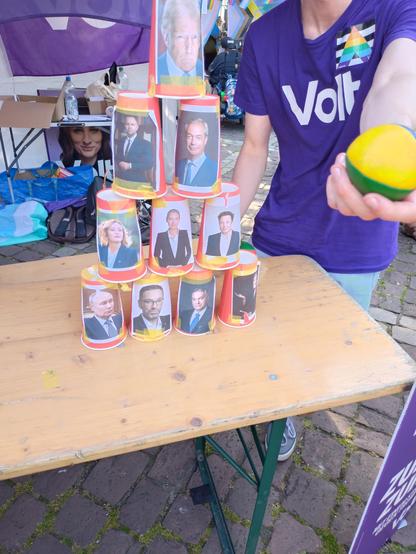 Pyramide aus umgedrehten pappbechern mit je 2 Löchern: 1 Loch zum Getränk einfüllen und eines, dessen Antlitz den Becher verschandelt. Unter Anderem : Putin, Orban, Musk, Weidel und dieser seltsame orange Irre den man aus den Nachrichten kennt