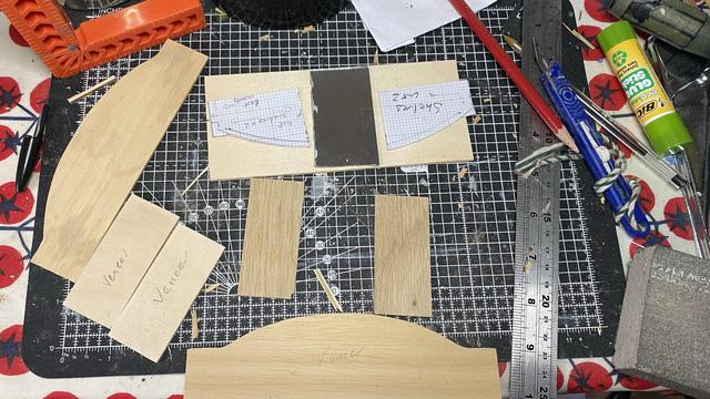 Pieces of wood and mirror on a cutting board with a pencil, ruler and craft knife visible. The base and top are curved. There’s two paper stencils saying ‘shelves cut 2’.
