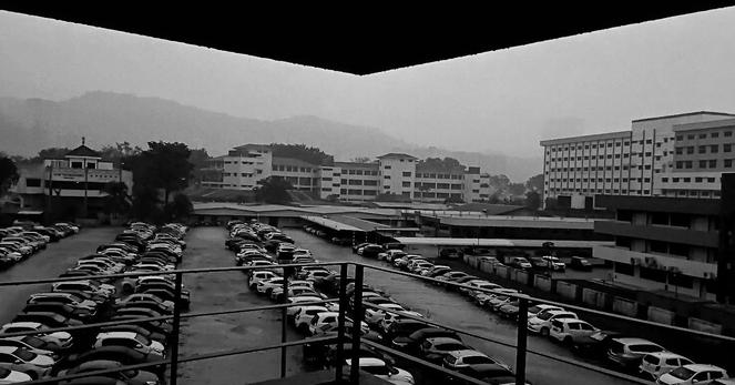 rain pours onto rows of cars in an open parking lot seen from above, framed by the ceiling and railing of a multi=story carpark... in black and white.