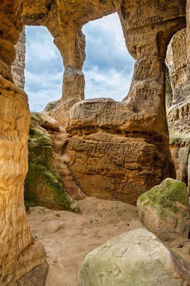 Sandstein Kammer mit zwei natürlichen Fenstern nach draußen. Auf dem Boden Sand. In den ganzen Sandstein Wänden sind Wörter eingraviert.