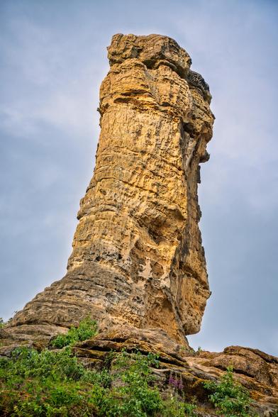 Sandstein Fels ragt empor. Im Hintergrund Wolkenhimmel. Unter dem Felsen Pflanzen, wie Fingerhut.