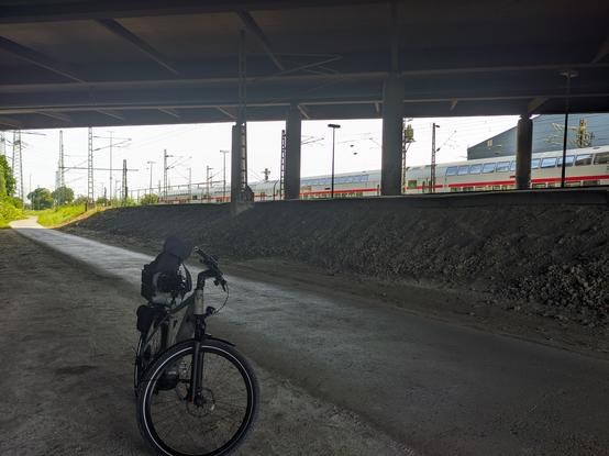 Ein Fahrrad ist unter einer Brücke auf einer asphaltierten Straße geparkt, mit einem Zug auf erhöhten Gleisen im Hintergrund.