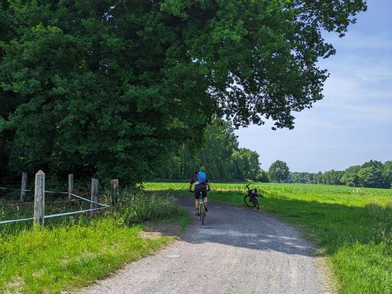 Eine Person fährt mit dem Fahrrad auf einem Kiesweg neben einem Feld und Bäumen, mit einem anderen Fahrrad parkt auf dem Gras in der Nähe unter einem klaren Himmel.