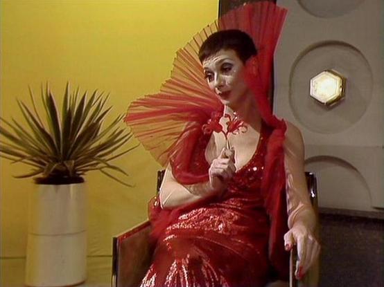 Claude 3.7 describes the image as: "The image shows a performer in a striking red sequined dress with a dramatic fan-like collar or accessory in matching red. Their theatrical makeup includes distinctive facial cosmetics that create a bold, stylized appearance. They're seated in what appears to be a retro-futuristic setting with a yellow wall, a potted plant, and geometric wall features including a hexagonal light fixture.

The scene has a theatrical, dramatic quality with the vibrant red costume contrasting against the yellow background. The styling and set design suggest this is from a science fiction production from the late 1970s or early 1980s, with the characteristic visual aesthetic of that era's television productions."

Blakes7Bot, version 3.9.30.