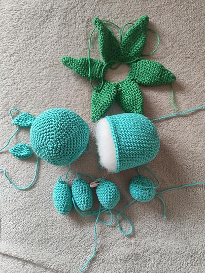 A collection of crocheted Bulbasaur (Bisasam) parts displayed on a light beige surface. The set includes two large blue-green body pieces—one of which is partially stuffed with white filling—six medium green leaf-like shapes arranged in a circle, four small turquoise ovals, and two smaller leaves. The yarn tails are visible, indicating the pieces are in progress and not yet assembled.