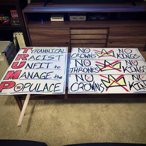 Three red, white, and blue protest signs lie on a living room coffee table: a large one that reads the name "TRUMP" as an acronym that stands for, "TYRANNICAL RACIST UNFIT to MANAGE the POPULACE," and the other three smaller ones that each read, "NO CROWNS | NO KINGS," "NO THRONES | NO KINGS," and another, "NO CROWNS | NO KINGS," each with a large sketched gold crown in the middle crossed out in red marker.