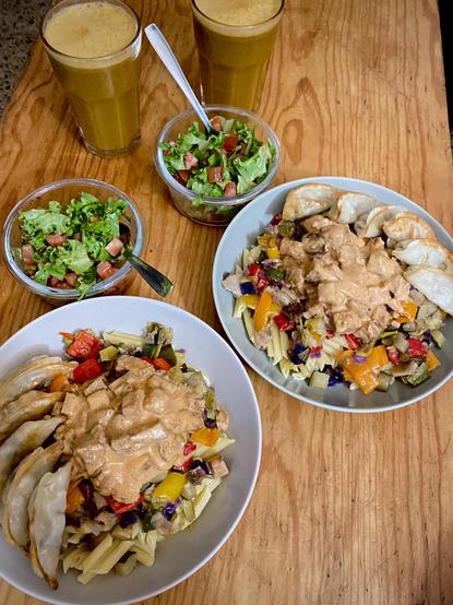 Two plates with veggie gyozas, pasta, veggie mix (leek, cucumber, green and red cabbage, and red, yellow and orange peppers), and seitan in a brown creamy sauce. Two little bowls of lettuce and tomato salad, seasoned with olive oil, apple cider vinegar and sea salt, and one with nori flakes and the other one with dried dill. Two big cups of fresh carrot, apple and cucumber juice.