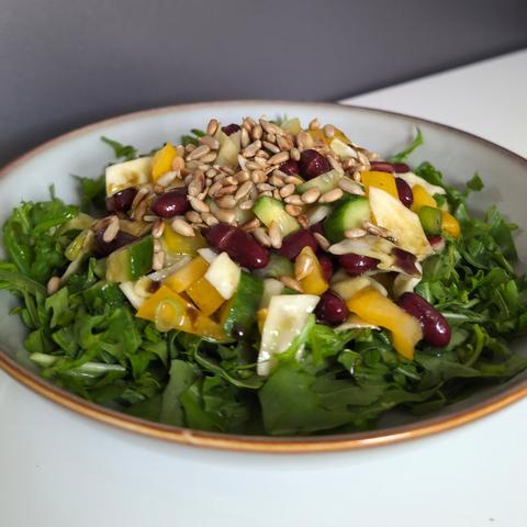 Picture of a kidney bean salad with cucumber, white cabbage and yellow bell pepper on a bed of cut arugula. The salad is garnished with roasted sunflower seeds and pumpkin seed oil. It is served on a gray plate. The background is a neutral white table.