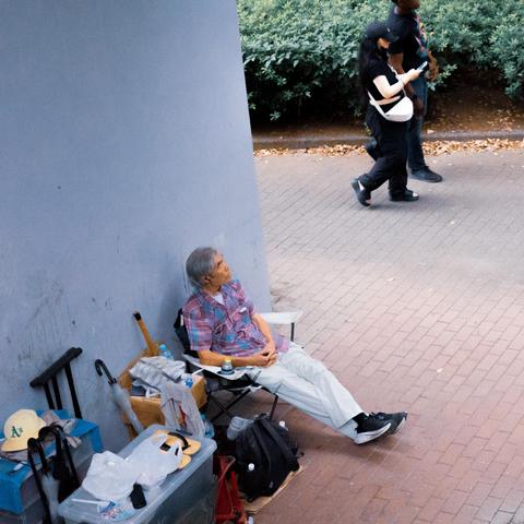 Elderly person sitting in a folding chair against a wall on a paved area. Surrounding them are personal belongings including a suitcase, a container, an umbrella, and a backpack. Two people walk on a path bordered by greenery in the background.