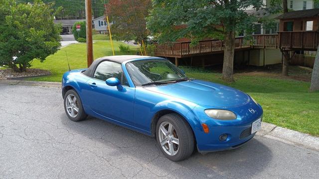 A convertible vibrant blue Miata with a black ragtop roof sitting in a very green suburban culd-de-sac.