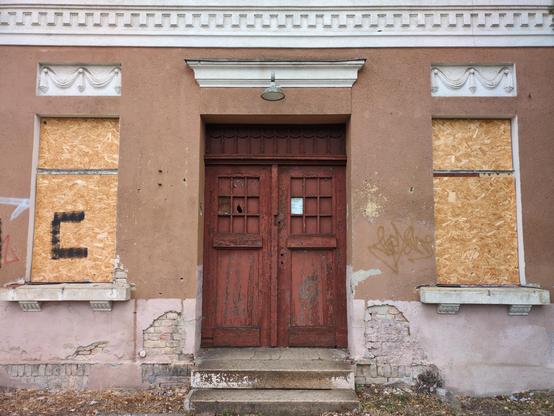 Front budynku dworca kolejowego. Pośrodku oszrupane, brązowe, podwójne drzwi. U góry drzwi mają kratownicę jak dla okien, ale wypełnioną drewnianymi płytami; klamkę usunięto. Po bokach dwa okna, zabite płytami OSB. Nad nimi ozdóbki i gzyms, koloru białego. Główna część ściany ma barwę jasnobrązową, przy ziemi zaś pasteloworóżową. Przy drzwiach odpadł tynk.