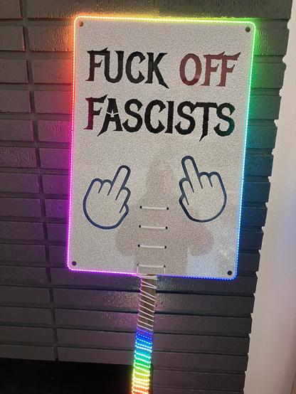 A sign cut out of holographic glitter acrylic says Fuck Off Fascists with two raised middle fingers.  There are LED COB lights lit up in a rainbow running around the edge of the sign and spiraling down the handle.