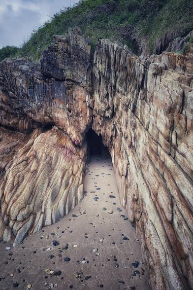 A narrow, dark cave entrance is framed by large, textured rock formations on a sandy and pebbled beach. The rocks on either side of the entrance show distinct horizontal layering and varying shades of brown, orange, and grey. Above the cave, a grassy, vegetated cliff rises, and the sky is overcast.