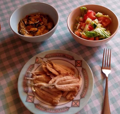 Un cuenco azul claro en el que se ven unos mejillones escabechados. Un cuenco beis con una ensalada de tomate, pepino, lechuga y cebolleta. Un plato de boquerones fritos.