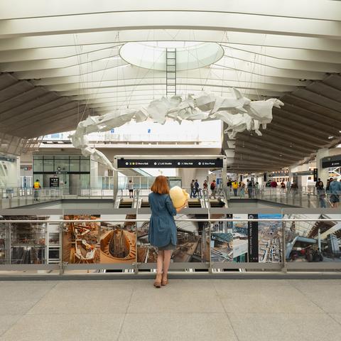 Photo of Lety and the Creature Mastodon stuffed toy standing on the second floor of the LAX/Metro Transit Center facing a giant, white origami-like sculpture suspended from the ceiling during a cloudy afternoon. From top to bottom, Lety is shown wearing a long-sleeve, thigh-length denim blue jacket, a gray bodycon dress, and suede pink boots. She's holding the Creature against her right bicep with her right hand, their backs to the camera as they look up at “the Distance of the Sun” sculpture by Glenn Kaino.