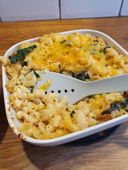 A square white baking dish containing baked macaroni and cheese with melted cheese on top, mixed with spinach. A serving spoon is partially immersed in the dish, and the dish is placed on a wooden surface.