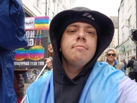A white transmasc wearing a black hoodie and black beanie. Round his shoulders is a bigender flag.
