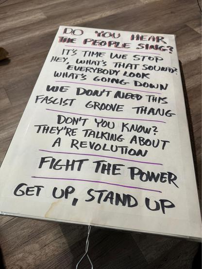 A handmade protest sign with song lyrics. From top to bottom: “Do you hear the people sing?”, “It’s time we stop, hey, what’s that sound? Everybody look what’s going down”, “We don’t need this fascist groove thang”, “Don’t you know? They’re talking about a revolution”, “Fight the power”, and “Get up, stand up”.