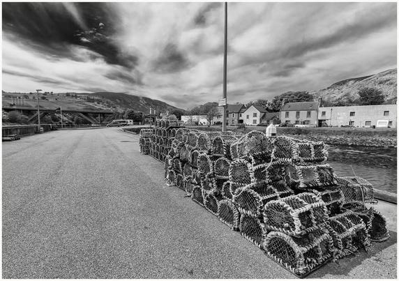 A black and white image of a harbor area featuring a stack of lobster traps on a paved road, with houses and hills visible in the background under a dramatic sky.