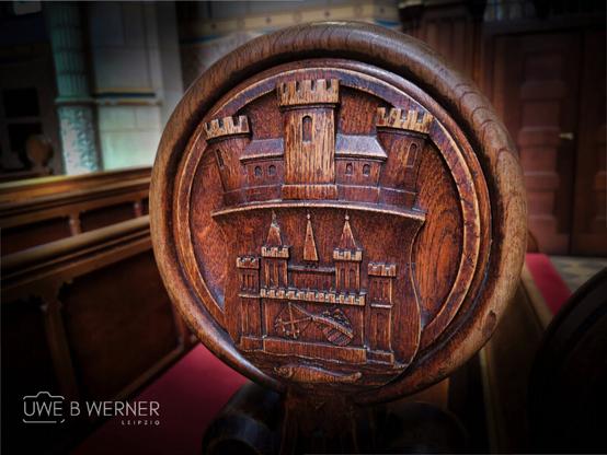 Detailaufnahme einer kunstvoll geschnitzten Holzlehne in der Schlosskirche Wittenberg, die das Wappen mit einer stilisierten Burg zeigt. Die Schnitzerei befindet sich an einem Kirchenstuhl und symbolisiert die historische Bedeutung der Kirche als Ursprungsort der Reformation.