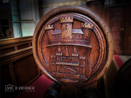 Detailaufnahme einer kunstvoll geschnitzten Holzlehne in der Schlosskirche Wittenberg, die das Wappen mit einer stilisierten Burg zeigt. Die Schnitzerei befindet sich an einem Kirchenstuhl und symbolisiert die historische Bedeutung der Kirche als Ursprungsort der Reformation.