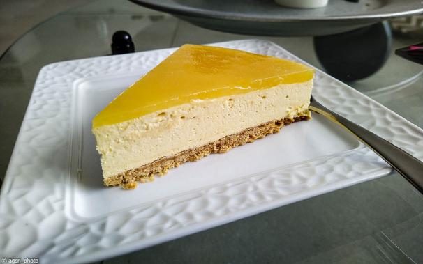 Auf einem weißen, quadratischen Kuchenteller liegt ein Stück Sanddorntorte, rechts daneben eine Kuchengabel.
Der Kuchen besteht aus drei Teilen: einem knusprigen braunen Boden, einer leichten Quark-Sahne-Füllung und einer gelben Schicht aus eingedicktem Sanddornsaft. /
A slice of sea buckthorn cake lies on a white, square cake plate, with a cake fork to the right. The cake consists of three parts: a crispy brown base, a light quark and cream filling, and a yellow layer of thickened sea buckthorn juice.