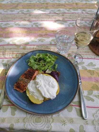 A table with a colorful tablecloth set with a blue plate. The table isnunder the trellis in the garden and the sunlight is playing on the tablecloth. On the plate you can see a baked potato with sour cream, a red onion, a green salad and a portion of salmon sprinkled with herbs. Above the plate are a glass of white wine and a glass of water.