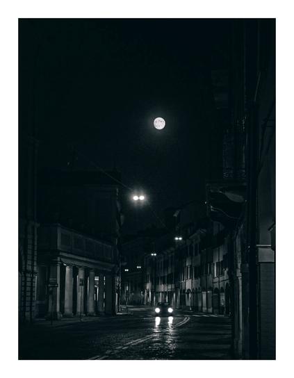 The photo is of a monochrome night shot of a street scene. A full moon hangs in the dark sky above a quiet, cobbled street lined with old, dimly lit buildings. There are streetlights that illuminate the road and lead your eye towards the centre of the photo. A single car's headlights are visible and is coming into the foreground.