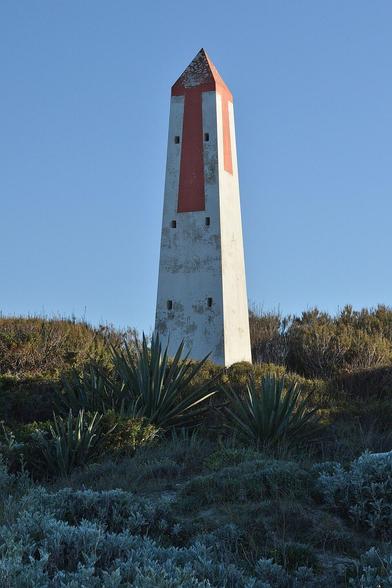 A photo of a white obelisk with red on it's tip and red and white on it's top half. There are six square holes visible on one of it's sides. It is on the top of a small hill covered in plants. The sky is clear and blue.