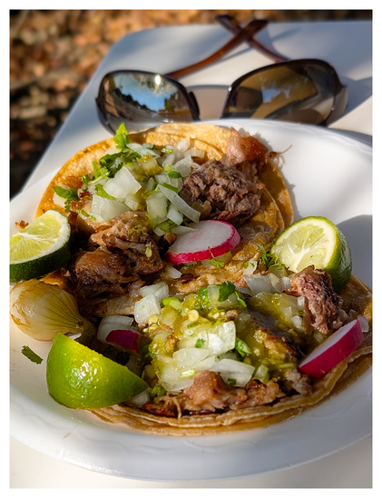 two carne asada tacos on double corn tortillas with onions, cilantro and salsa verde on a white disposable plate with sliced radishes, a grilled spring onion and lime wedges. the plate sits on a white plastic picnic table, in dappled light with a pair of sunglasses that reflect the scene.