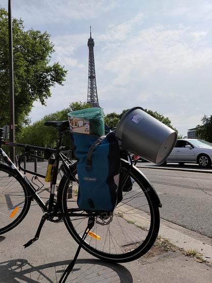 Photographie d'un vélo de côté devant la Tour Eiffel. Sur le porte-bagages arrière du vélo, une grande sacoche bleue ouverte dont dépasse en haut un rouleau de papier à bulles. Dépassant du porte-bagages arrière, un seau gris vide est fixé à la sacoche par son anse.