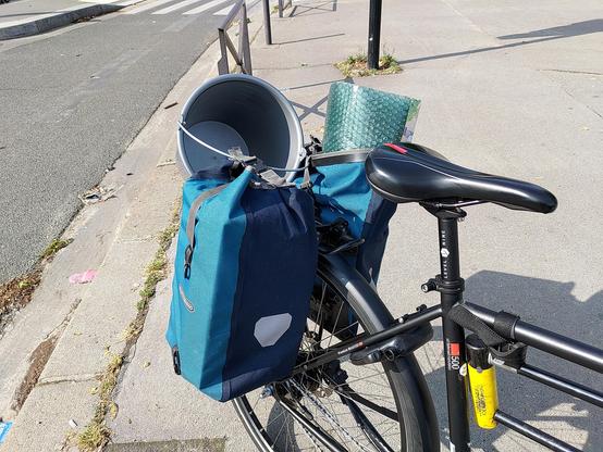 Photographie de l'arrière d'un vélo. Sur le porte-bagages sont fixées deux grosses sacoches noires. La première ferme tout juste tellement elle est remplie. La seconde est ouverte et une rouleau de papier à bulles en dépasse. Un seau gris vide est fixé aux sacoches par son anse.