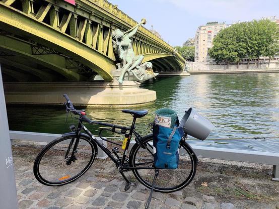 Photographie d'un vélo de côté sur un quai et devant un pont traversant la Seine. Sur le porte-bagages arrière du vélo, une grande sacoche bleue ouverte dont dépasse en haut un rouleau de papier à bulles. Dépassant du porte-bagages arrière, un seau gris vide est fixé à la sacoche par son anse.