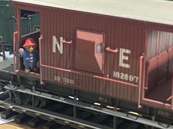 Close up of a model brake van in a dirty dark red colour. In the doorway leaning out and waving is Paddington Bear wearing his trademark red hat and blue coat.