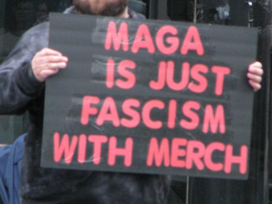 A protest sign. Red lettering on a black background which reads "MAGA is just fascism with merch"