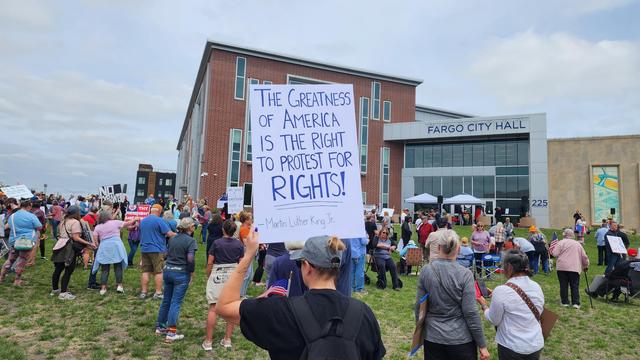 The image depicts a protest gathering in front of Fargo City Hall. A large crowd is visible, with many individuals holding signs. The most prominent sign in the foreground reads, "The greatness of America is the right to protest for rights!" attributed to Martin Luther King Jr. The crowd is diverse, with people of various ages and attire, some wearing casual clothing and others in more formal attire. The setting is outdoors, with a grassy area in front of the building, and the sky is overcast. The building itself is a modern structure with a brick facade and large windows, labeled "Fargo City Hall" and numbered "225." The atmosphere appears peaceful, with people standing and sitting, some holding American flags. The scene conveys a sense of civic engagement and the exercise of First Amendment rights.
