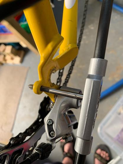 The rear drop out of a yellow bike, pictured from above. The grey rear derailleur is attached below the drop out. Just behind the drop out a frame tap is inserted into the boss for attaching accessories such as mudguards or a rear rack. The tap looks like a drill bit with threads. At the other end of the tap is a long silver and black handle for turning the tap.