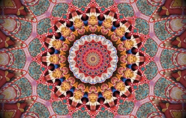 A radial kaleidoscope made from a photo of embroidered textiles in pink, red, teal, and yellow. The image is flower-like and has the detail of stitches and fringe here and there.