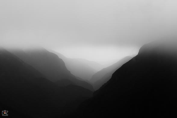 Black and white photograph of a V-shaped valley with various levels of silhouetted mountains or mountain slopes, which become lighter from the front to the back (black to light gray). 

The sky is gray without structure and partially covers the mountains as fog.