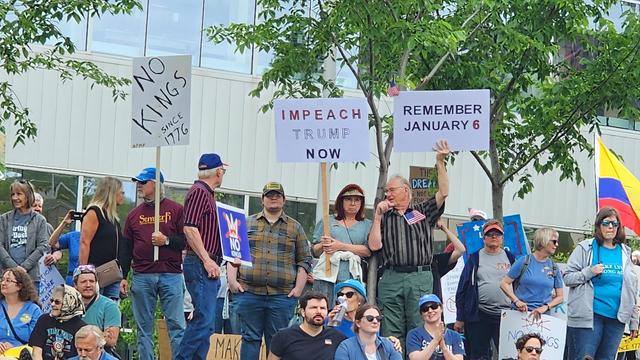 The image depicts a group of people gathered for a protest. They are standing and sitting in front of a building with large windows. The protesters are holding various signs with messages such as "NO KINGS SINCE 1776," "IMPEACH TRUMP NOW," and "REMEMBER JANUARY 6." The signs are held up by individuals of different ages and genders, wearing casual clothing. Some signs have additional text, including "MAKE AMERICA GREAT AGAIN" and "NO KINGS." A Colombian flag is visible in the background, adding to the diversity of the crowd. The setting appears to be outdoors, with trees and a clear sky visible. The protesters seem to be engaged in a peaceful demonstration, with some individuals looking at their signs and others conversing with each other.