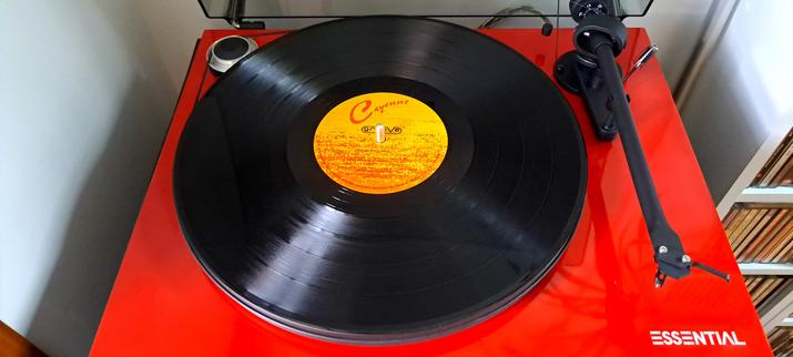 A shiny vinyl LP record with a bright orange/red label sits on a gloss red turntable, waiting to be cued up for an appreciative audience of two.