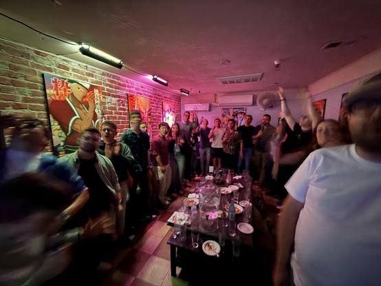 A group of about 30 people stands in a karaoke room, a low table in the middle and brick walls behind them. The scene is blurred with people, singing, and moving to the music.