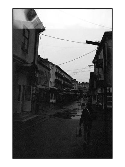 The image depicts a narrow street in an urban setting, captured in black and white, which adds a timeless quality to the scene. The street is flanked by multi-story buildings with a mix of residential and commercial architecture, featuring balconies and awnings. The buildings are closely packed, with visible windows and doors, and some have signs, suggesting shops or businesses. Overhead, a network of power lines crisscrosses the sky, adding to the urban feel. The street is wet, indicating recent rain, with puddles reflecting the buildings and sky. A solitary figure is seen walking away from the camera, carrying a bag, adding a sense of scale and life to the scene. The overall atmosphere is quiet and somewhat melancholic, with the overcast sky contributing to the subdued lighting.