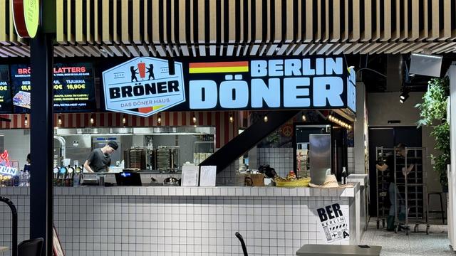 Image of a food stall named "Berlin Döner" with a large illuminated sign. The stall features a menu board displaying various items and prices. A chef is seen working behind the counter, with kitchen equipment visible. The decor includes modern design elements and